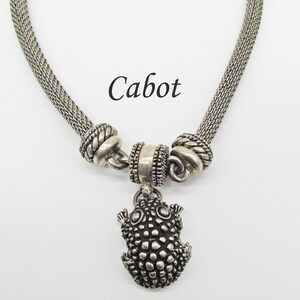May include: Silver chain necklace with a frog pendant. The chain is made of a mesh design and has two decorative beads. The pendant is a detailed frog with a textured surface.