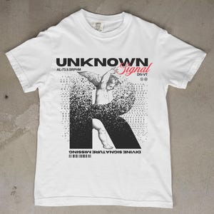 Könnte beinhalten: Weißes T-Shirt mit den Worten "UNKNOWN Signal" in Schwarz und Rot. Das Shirt zeigt eine Grafik eines Engels und den Buchstaben "R". Der Text "DIVINE SIGNATURE MISSING" ist unten aufgedruckt.