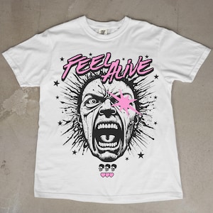 Könnte beinhalten: Weißes T-Shirt mit einem schwarz-rosa Grafikdesign. Das Design zeigt ein schreiendes Gesicht mit den Worten "FEEL ALIVE" darüber, sowie Sterne und Herzmotive.