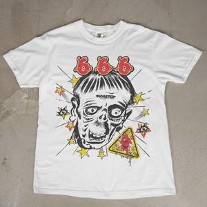 Pode incluir: Camiseta branca com um gráfico de um rosto semelhante a um zumbi, o número 666 acima da cabeça, estrelas amarelas, olhos e um sinal de aviso com uma figura vermelha. A palavra "MOMSTER" é impressa abaixo da cabeça.
