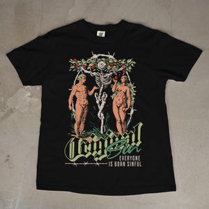 Könnte beinhalten: Schwarzes T-Shirt mit einem Grafikdesign, das ein Skelett, zwei nackte Figuren und den Text "Legal Sin" zeigt. Das Design enthält grüne und rote Akzente mit dem Text "Everyone is Born Sinful".