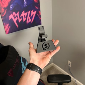 May include: A black phone stand with a small 'S' logo on the front. The stand is made of plastic and has a triangular shape.