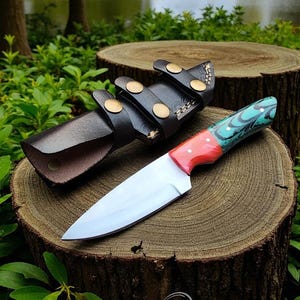 May include: A handmade knife with a polished steel blade and a handle featuring red, teal, and black patterns. The knife rests on a wooden surface next to a black leather sheath with brass accents. The knife is approximately 10 inches long.