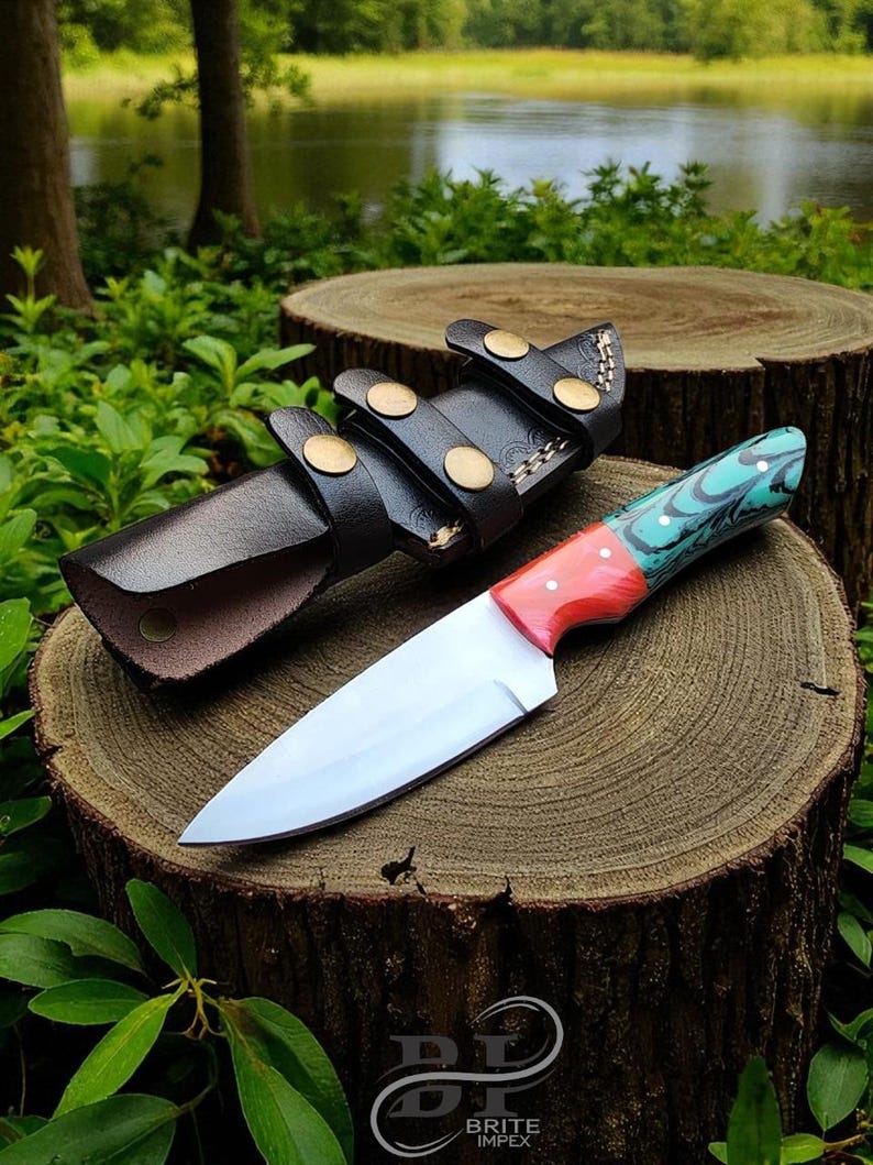 May include: A handmade knife with a polished steel blade and a handle featuring red, teal, and black patterns. The knife rests on a wooden surface next to a black leather sheath with brass accents. The knife is approximately 10 inches long.