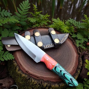 May include: A handcrafted knife with a silver blade and a handle featuring coral and turquoise resin. The knife rests on a wooden surface with its leather sheath, secured by black straps and gold-colored rivets. The knife is surrounded by greenery.