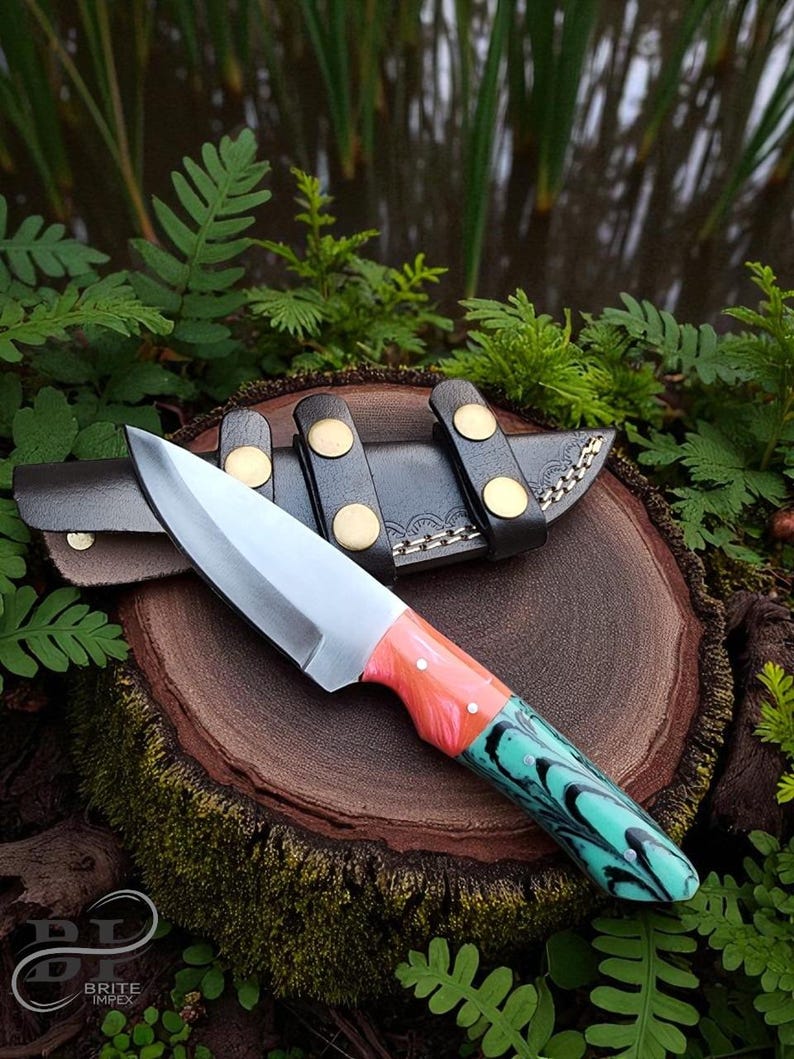 May include: A handcrafted knife with a silver blade and a handle featuring coral and turquoise resin. The knife rests on a wooden surface with its leather sheath, secured by black straps and gold-colored rivets. The knife is surrounded by greenery.