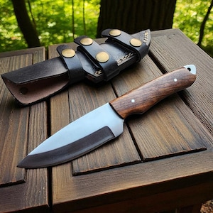 May include: A handcrafted hunting knife with a wooden handle and a stainless steel blade, resting on a wooden surface. The knife is accompanied by a black leather sheath with brass-colored button closures. The background features a blurred green forest.