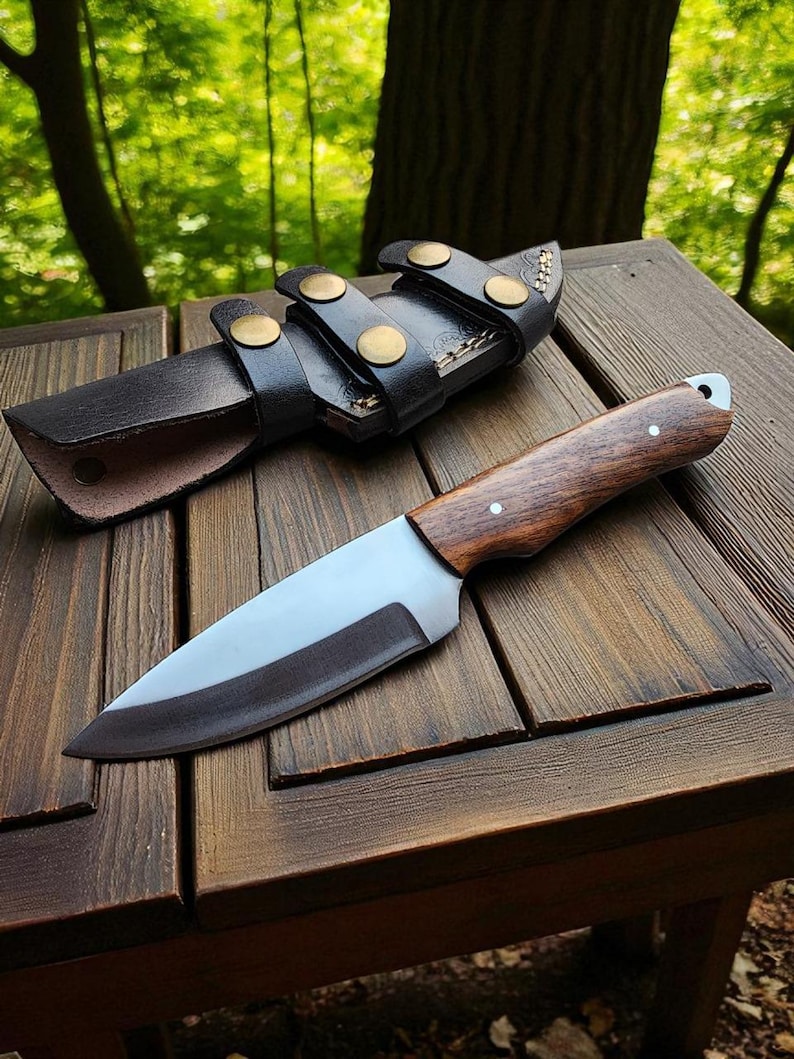 May include: A handcrafted hunting knife with a wooden handle and a stainless steel blade, resting on a wooden surface. The knife is accompanied by a black leather sheath with brass-colored button closures. The background features a blurred green forest.