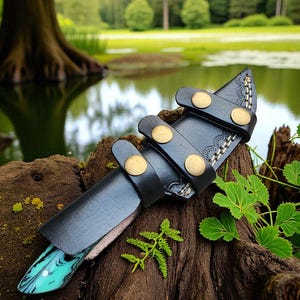 May include: A handcrafted knife with a black leather sheath. The knife handle has a turquoise and white marbled design. The sheath features brass rivets and decorative stitching. The knife is displayed on a piece of wood with a natural background.