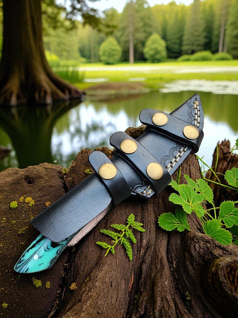 May include: A handcrafted knife with a black leather sheath. The knife handle has a turquoise and white marbled design. The sheath features brass rivets and decorative stitching. The knife is displayed on a piece of wood with a natural background.