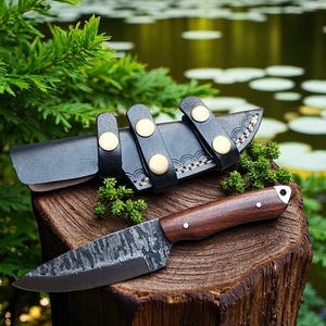 May include: A handcrafted knife with a dark, textured blade and a wooden handle rests on a tree stump. The knife is accompanied by a black leather sheath with gold-colored button closures. The scene is set outdoors with greenery and water in the background.