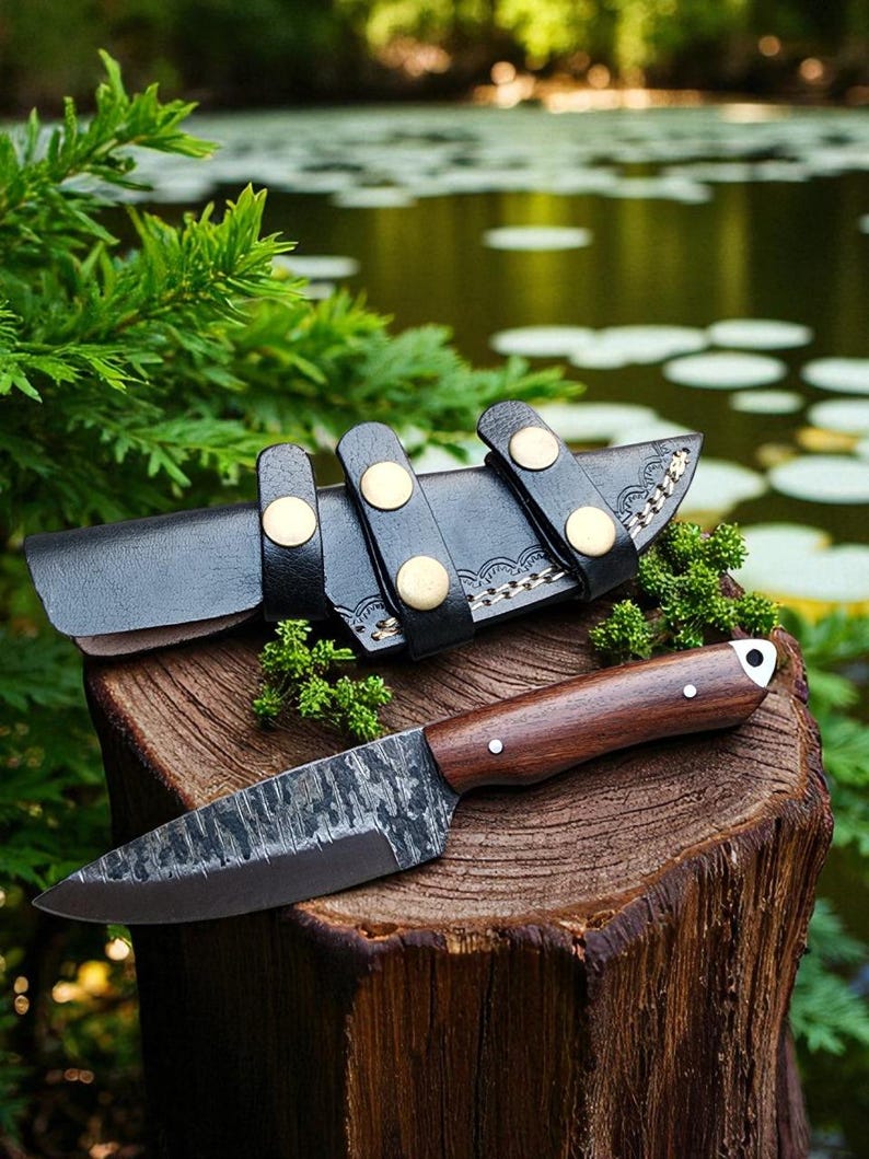 May include: A handcrafted knife with a dark, textured blade and a wooden handle rests on a tree stump. The knife is accompanied by a black leather sheath with gold-colored button closures. The scene is set outdoors with greenery and water in the background.