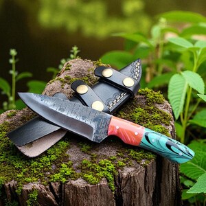 May include: A handcrafted knife with a patterned blade and a colorful handle featuring red, orange, and teal hues. The knife rests on a moss-covered tree stump, accompanied by a black leather sheath with brass accents. The background is a blurred view of greenery.