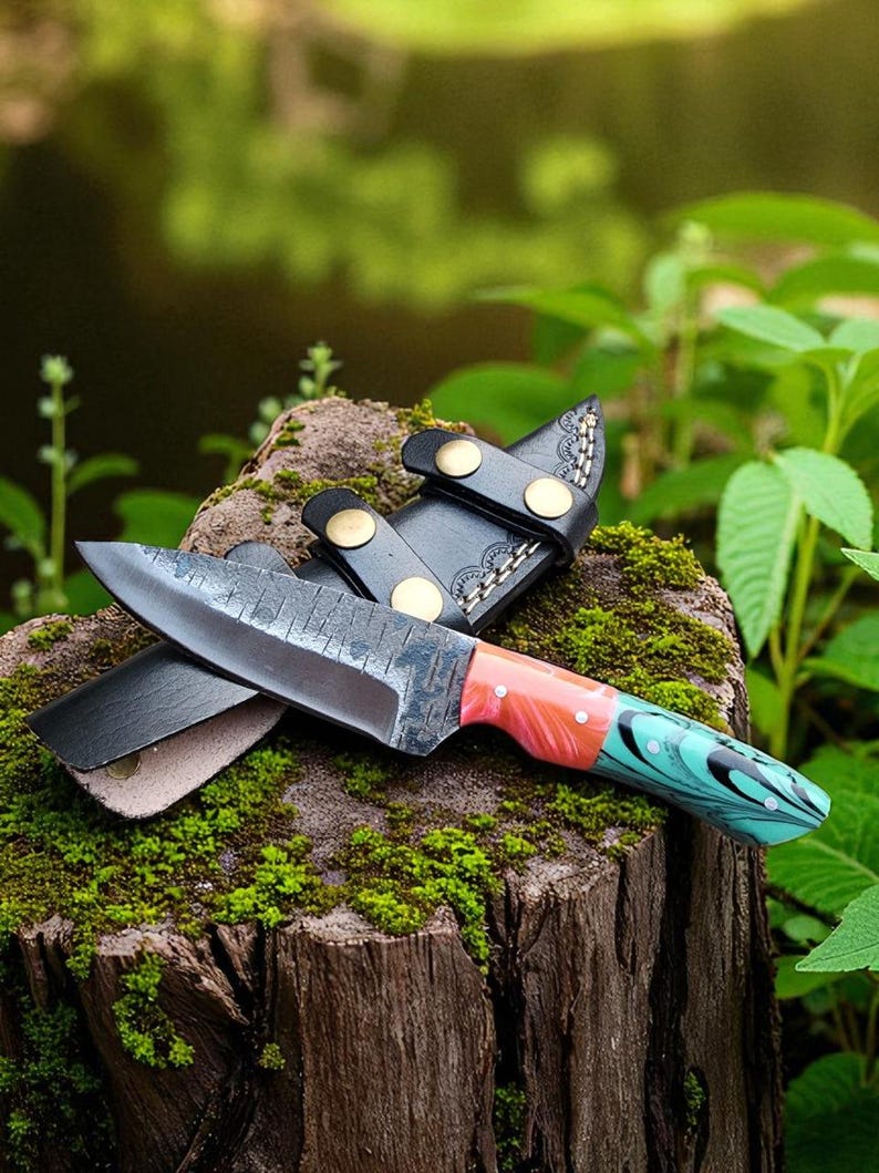 May include: A handcrafted knife with a patterned blade and a colorful handle featuring red, orange, and teal hues. The knife rests on a moss-covered tree stump, accompanied by a black leather sheath with brass accents. The background is a blurred view of greenery.