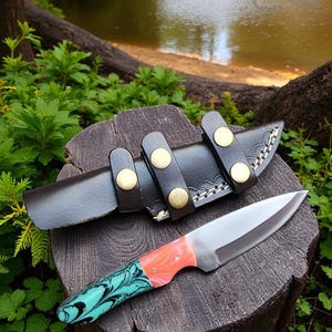 May include: A handmade knife with a stainless steel blade and a handle featuring a teal and coral swirl design. The knife rests on a wooden stump next to a black leather sheath with brass button closures.