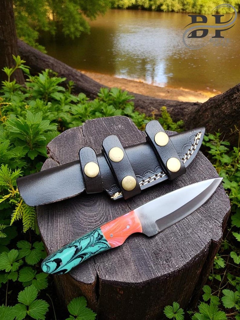 May include: A handmade knife with a stainless steel blade and a handle featuring a teal and coral swirl design. The knife rests on a wooden stump next to a black leather sheath with brass button closures.
