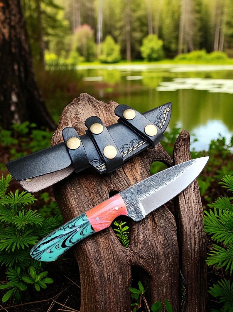 May include: A handcrafted knife with a patterned blade and a handle featuring shades of teal, pink, and orange. The knife rests on a piece of wood, with a black leather sheath secured by brass-colored studs. The background shows a lake and greenery.