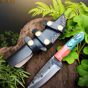 May include: A handcrafted knife with a textured blade and a colorful handle. The handle features red, teal, and white accents. The knife is paired with a black leather sheath secured by brass-colored studs. The knife and sheath rest on a wooden surface.