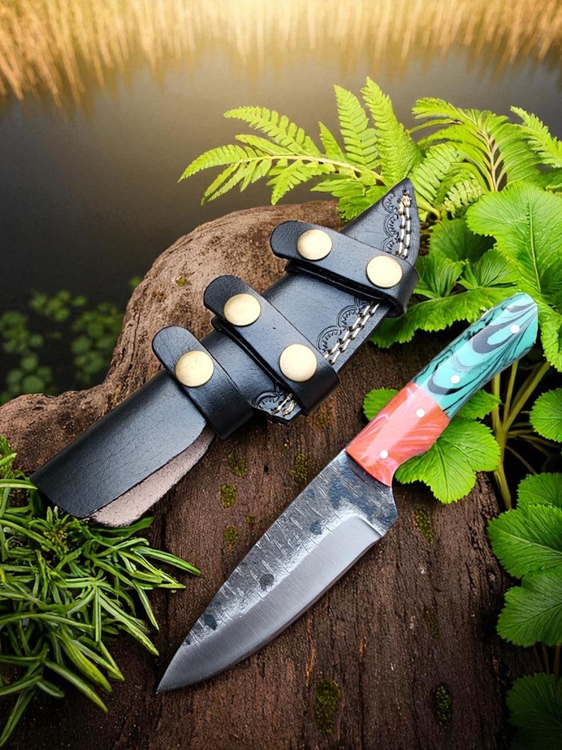 May include: A handcrafted knife with a textured blade and a colorful handle. The handle features red, teal, and white accents. The knife is paired with a black leather sheath secured by brass-colored studs. The knife and sheath rest on a wooden surface.