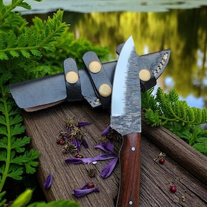 May include: A handcrafted knife with a wooden handle and a textured blade, resting on a wooden surface. The knife is accompanied by a black leather sheath with gold-colored rivets. Purple flower petals and greenery surround the knife, suggesting an outdoor setting.
