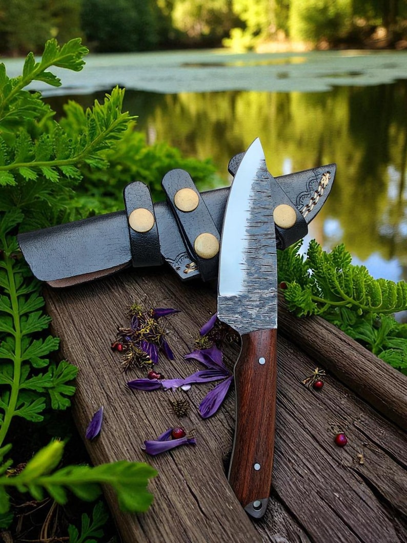 May include: A handcrafted knife with a wooden handle and a textured blade, resting on a wooden surface. The knife is accompanied by a black leather sheath with gold-colored rivets. Purple flower petals and greenery surround the knife, suggesting an outdoor setting.