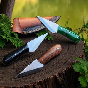 May include: Three handcrafted knives with different handle materials: green, black, and wood. Each knife has a silver blade and is displayed on a wooden surface. A brown leather sheath is visible in the background, suggesting outdoor or hunting use.