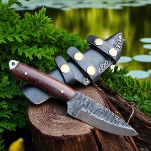 May include: A handcrafted knife with a dark wooden handle and a textured blade, resting on a wooden stump. The knife is accompanied by a black leather sheath with brass-colored fasteners. The scene is set outdoors with greenery and water in the background.
