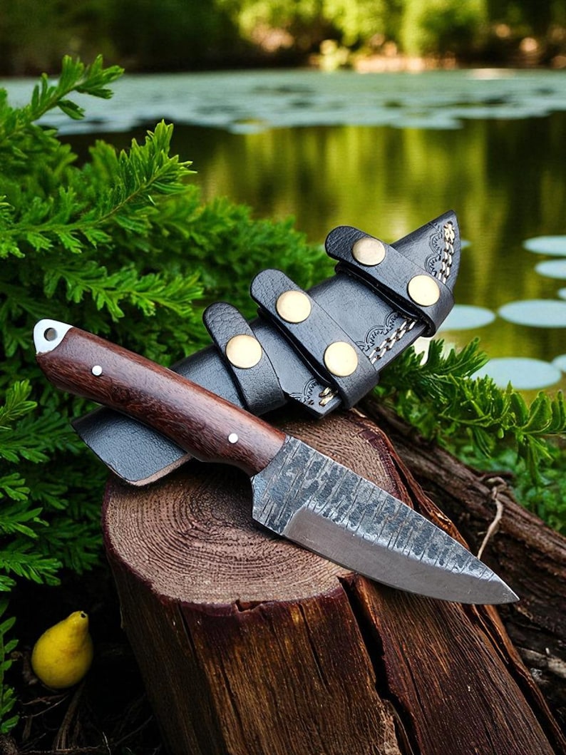 May include: A handcrafted knife with a dark wooden handle and a textured blade, resting on a wooden stump. The knife is accompanied by a black leather sheath with brass-colored fasteners. The scene is set outdoors with greenery and water in the background.