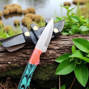 May include: A stainless steel knife with a colorful handle featuring shades of orange, pink, and teal. The knife is resting on a wooden log next to a black leather sheath with gold-colored rivets. The background includes water and green foliage.