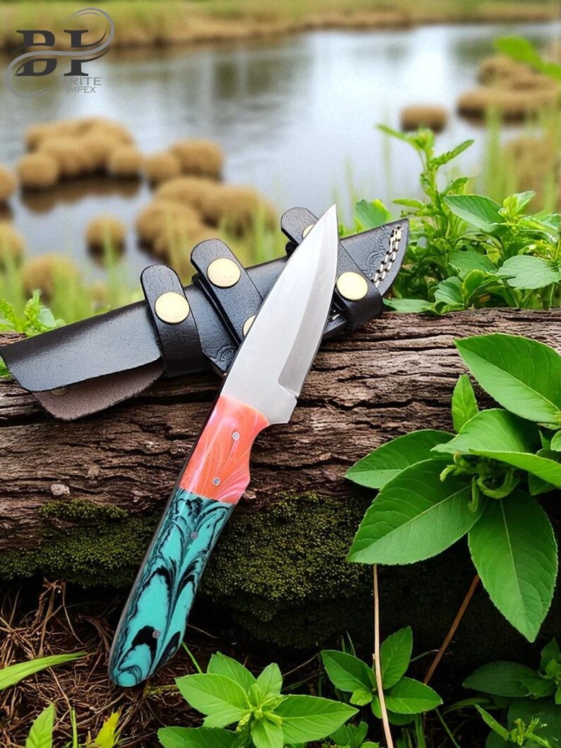 May include: A stainless steel knife with a colorful handle featuring shades of orange, pink, and teal. The knife is resting on a wooden log next to a black leather sheath with gold-colored rivets. The background includes water and green foliage.