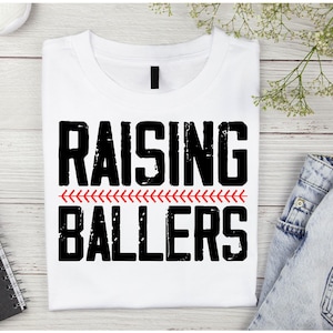 May include: White t-shirt with black text that reads "Raising Ballers" with a red baseball stitch design underneath the text.
