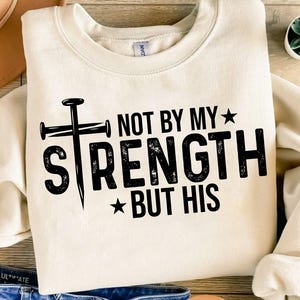 Puede incluir: Sudadera crema con el texto negro "NOT BY MY STRENGTH BUT HIS". El diseño incluye una cruz hecha de clavos y estrellas. La sudadera está doblada sobre una superficie de madera.