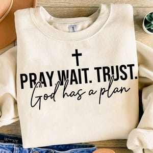 May include: Cream-colored sweatshirt with the text "PRAY WAIT. TRUST. God has a plan" in black, with a cross above the text. The sweatshirt is folded on a wooden surface, with a hat and jeans visible.