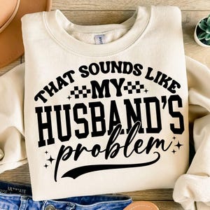 Może przedstawiać: Kremowa bluza z napisem "THAT SOUNDS LIKE MY HUSBAND'S problem" w kolorze czarnym. Tekst jest pogrubioną, zabawną czcionką. Bluza jest złożona na drewnianej powierzchni.