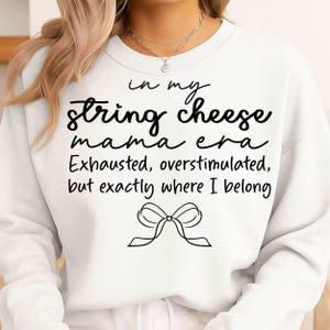 May include: White sweatshirt featuring the text "in my string cheese mama era Exhausted, overstimulated, but exactly where I belong" in black script. A black bow graphic is at the bottom. The sweatshirt is a casual style.