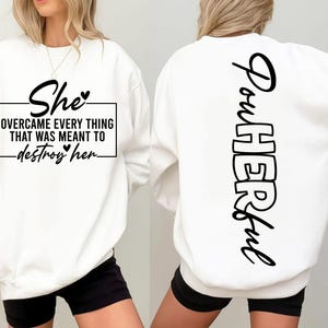 Könnte beinhalten: Weißes Sweatshirt mit schwarzem Text. Auf der Vorderseite steht: „She overcame everything that was meant to destroy her“. Auf der Rückseite ist das Wort „Powerful“ vertikal in Schwarz gedruckt.