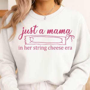 Könnte beinhalten: Weißes Sweatshirt mit dem pinkfarbenen Text "just a mama in her string cheese era". Das Design beinhaltet eine rosa Käsesnack-Grafik. Das Sweatshirt ist ein lässiger, bequemer Stil.