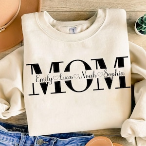 May include: A cream-colored sweatshirt with the word "MOM" in large black letters. The letters are decorated with small hearts and the names "Emily", "Lucas", "Noah", and "Sophia" written underneath.