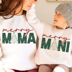 May include: White sweatshirts with matching "Merry Mama" and "Merry Mini" Christmas designs in green and red. The "Mama" sweatshirt features a Christmas tree graphic. The "Mini" sweatshirt is worn by a child wearing a gray beanie.