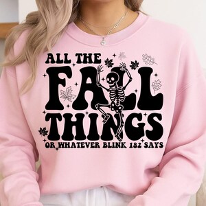 May include: Pink sweatshirt with the text "ALL THE FALL THINGS" in black, featuring a skeleton holding leaves. The phrase "OR WHATEVER BLINK 182 SAYS" is below the main text. Autumn-themed apparel.