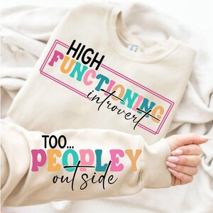Könnte beinhalten: Cremefarbenes Sweatshirt mit dem Text "HIGH FUNCTIONING introvert" in einem farbenfrohen rechteckigen Design. Der Ärmel zeigt den Text "TOO... PEOPLE outside". Das Sweatshirt ist gefaltet.