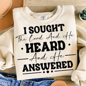 Puede incluir: Sudadera color crema con el texto negro "I SOUGHT The Lord And He HEARD And He ANSWERED". La sudadera está doblada, con un sombrero y unos vaqueros visibles en el fondo. Una pequeña planta en maceta también está en el fondo.