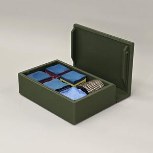 May include: An olive green box with a hinged lid, open to reveal several small, colorful items. Inside are blue and yellow squares, and a roll of silver and gold items. The box is on a light gray surface.