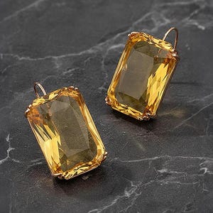 May include: Two rectangular earrings featuring yellow gemstones in gold-coloured settings. The earrings have a hook design and are set against a dark, marbled surface. The gemstones are faceted, reflecting light.