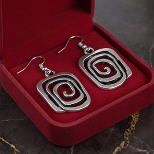 May include: Silver-coloured earrings featuring a square spiral design. The earrings are presented in a red velvet jewellery box. The spiral design is a key feature, creating a unique look. The hooks are silver.