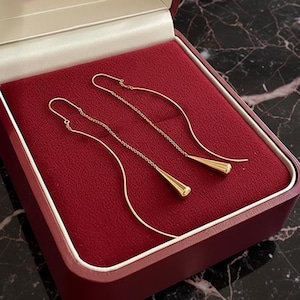 May include: Gold-coloured drop earrings presented in a red jewellery box. The earrings feature a delicate chain design with a teardrop-shaped pendant. The box is open, revealing a soft, velvety interior.