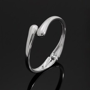 May include: A polished silver bangle bracelet with a distinctive open design. The bracelet has a smooth, curved band that encircles the wrist, with two teardrop-shaped ends that meet at the top. The bracelet has a clasp closure.
