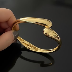 May include: A gold-coloured, open-ended cuff bracelet. The bracelet has a smooth, polished finish and a curved design. The bracelet is held by a hand with dark grey nail polish. The background is black.