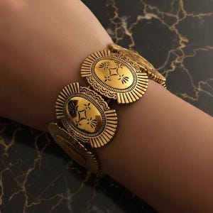 May include: A gold-coloured bracelet with oval-shaped medallions, each adorned with a floral pattern. The bracelet is made up of several linked sections, each with a detailed design and a sunburst edge. The jewellery is displayed on a wrist, highlighting its intricate design.