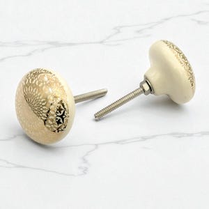 May include: Two cream ceramic cabinet knobs with a gold floral pattern. Each knob features a silver threaded screw for fitting. The knobs are round and have a decorative design on the front.
